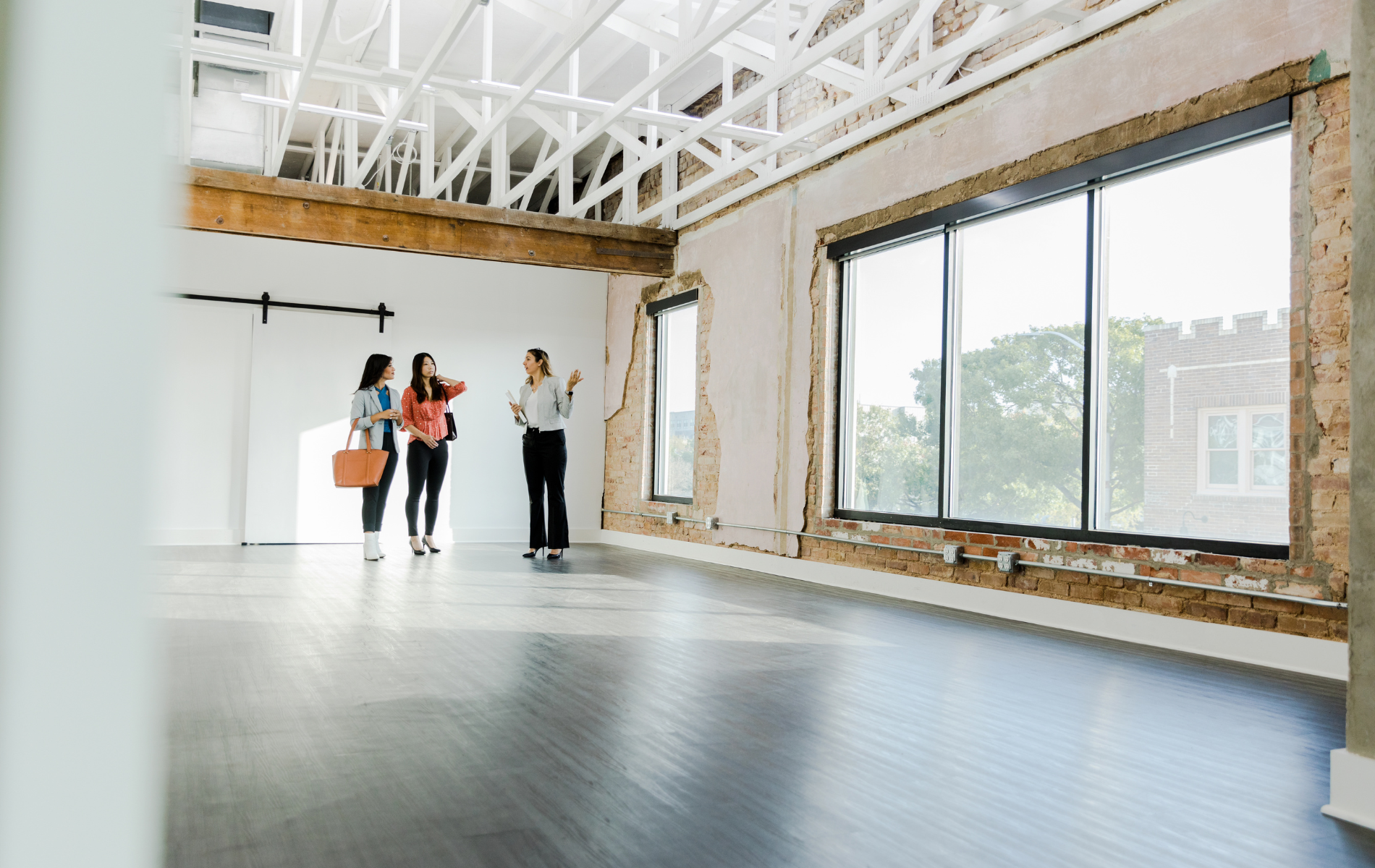 Three people in an empty industrial space, looking at windows. Exposed beams and brick walls.
