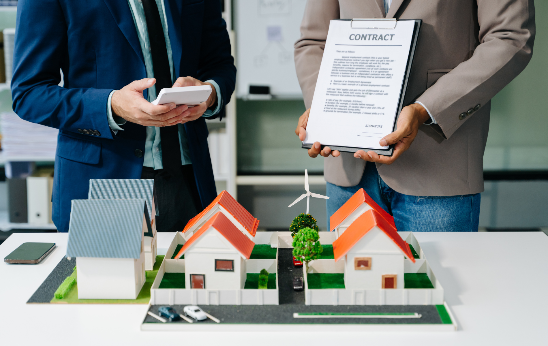 Two men review a contract and model houses on a table. One holds a calculator.