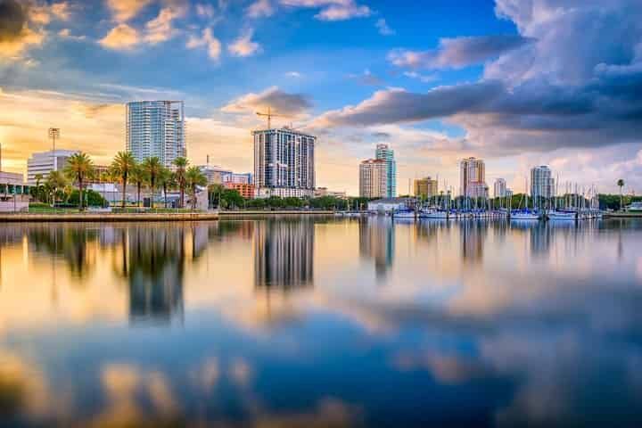City skyline reflected in water at sunset, with boats, palm trees, and cloudy sky.