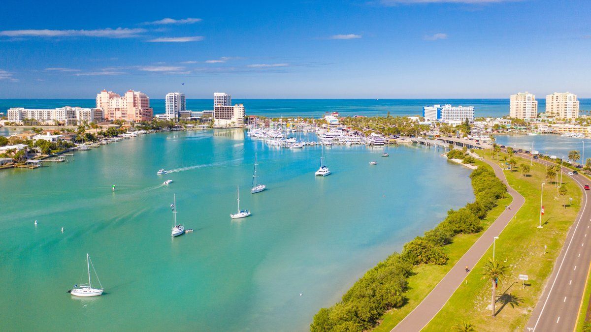 Boats sail in a turquoise bay near a city with tall buildings under a blue sky.