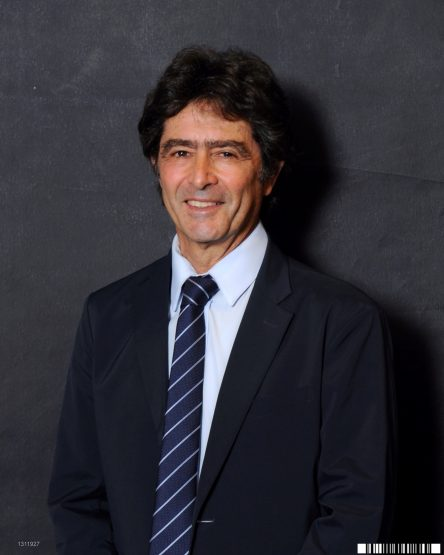 Man in suit smiles. Dark hair, blue tie, against dark background.