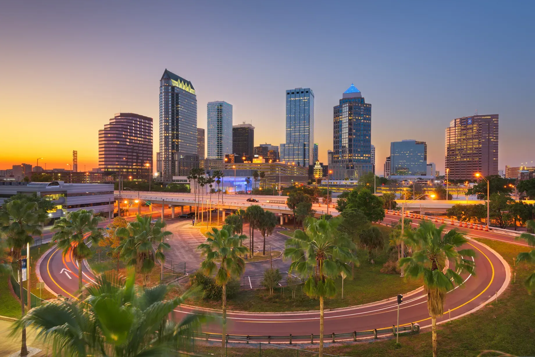 Sunset skyline over a city park with palm trees, curved roads, and illuminated high-rise buildings