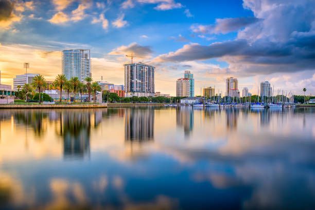 City skyline reflecting in calm water at sunset, with boats and palm trees.