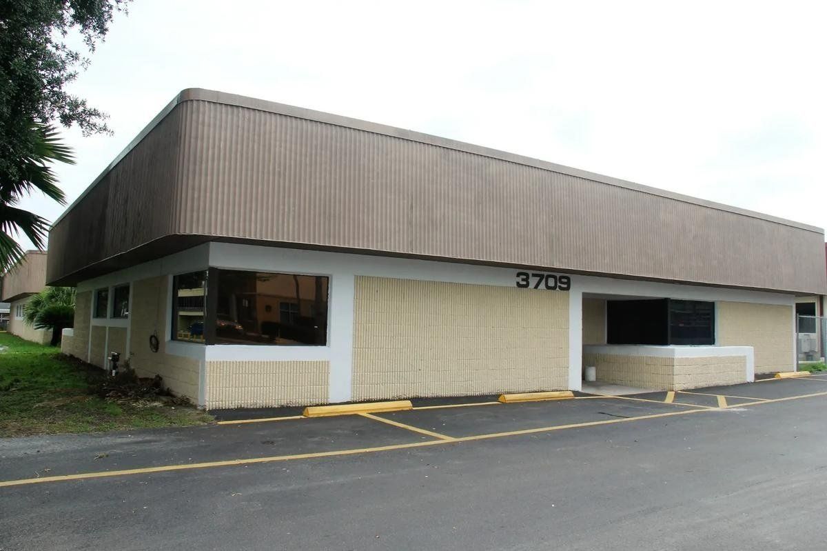 Low-rise beige commercial building with brown awning, windows, and parking lot in front