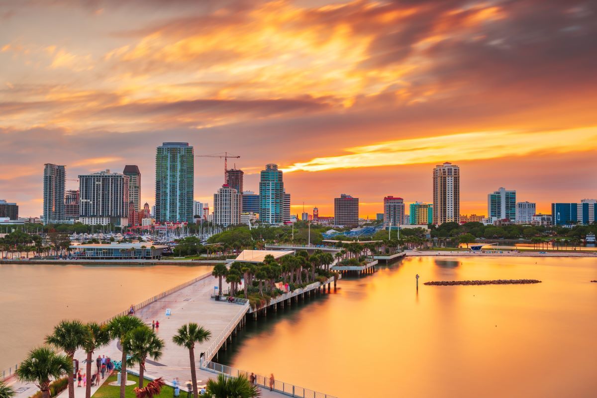 Skyline of St. Petersburg, Florida at sunset, featuring high-rise buildings, pier, and orange-yellow sky reflecting in water.