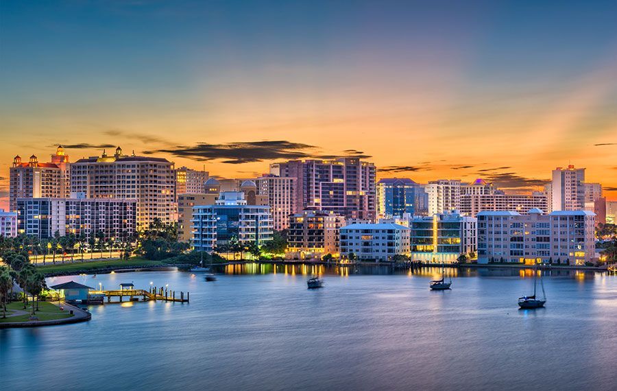 City skyline at sunset, waterfront buildings, boats in the water, vibrant orange and blue sky.
