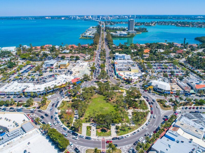 Aerial view of a coastal town with a roundabout, bridge, buildings, and clear blue water.