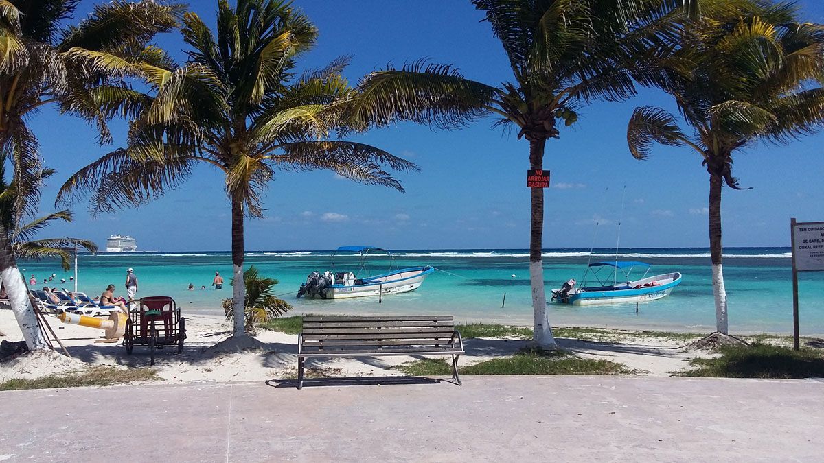 Beach scene: Palm trees, turquoise water, boats, people, and white sand. Blue sky.