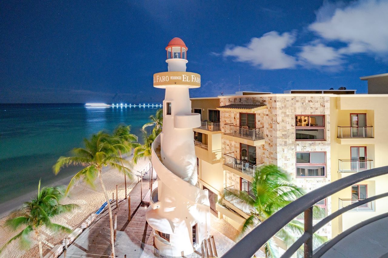 Beachfront hotel with a lighthouse, palm trees, and ocean under a night sky.