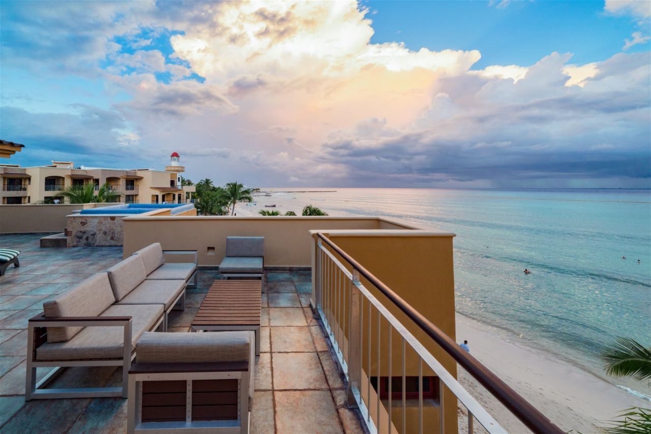 Rooftop lounge overlooking ocean. Beige sofas and tables, sandy beach, cloudy sky.