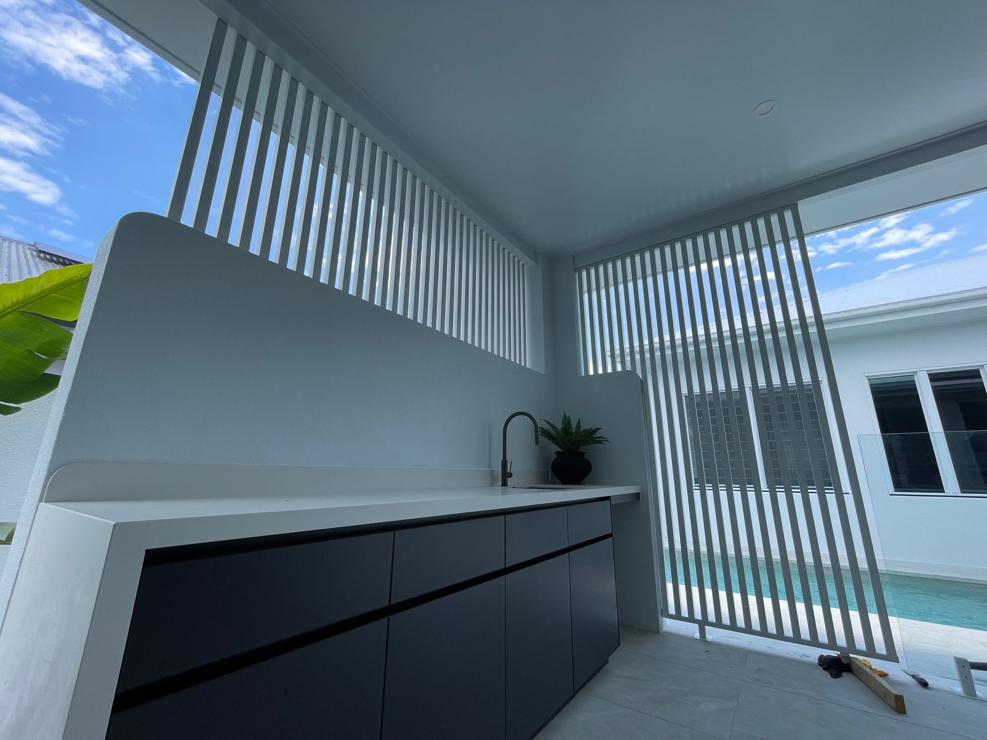 Outdoor kitchen with dark cabinets, white countertop, and slatted screen, with blue sky and pool visible — Lifestyle Aluminium Lattice in West End, QLD