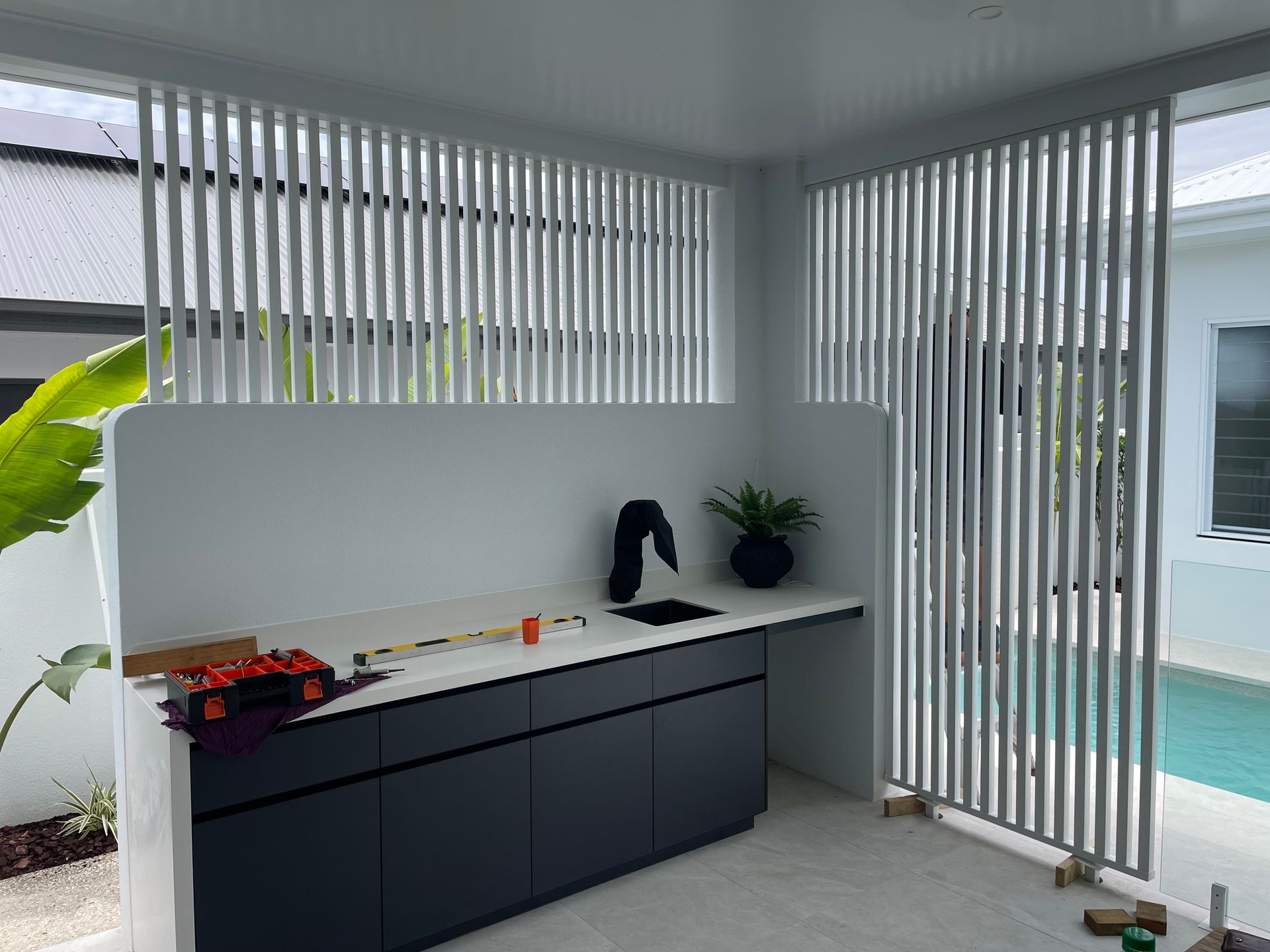 Outdoor kitchen with black cabinets, white countertop, vertical slat screens, and a pool in the background — Lifestyle Aluminium Lattice in West End, QLD