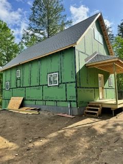 Green house under construction with unfinished siding, front porch, and tall trees in the background
