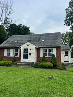 Small beige house with white trim, dark roof, and a large front lawn under cloudy skies