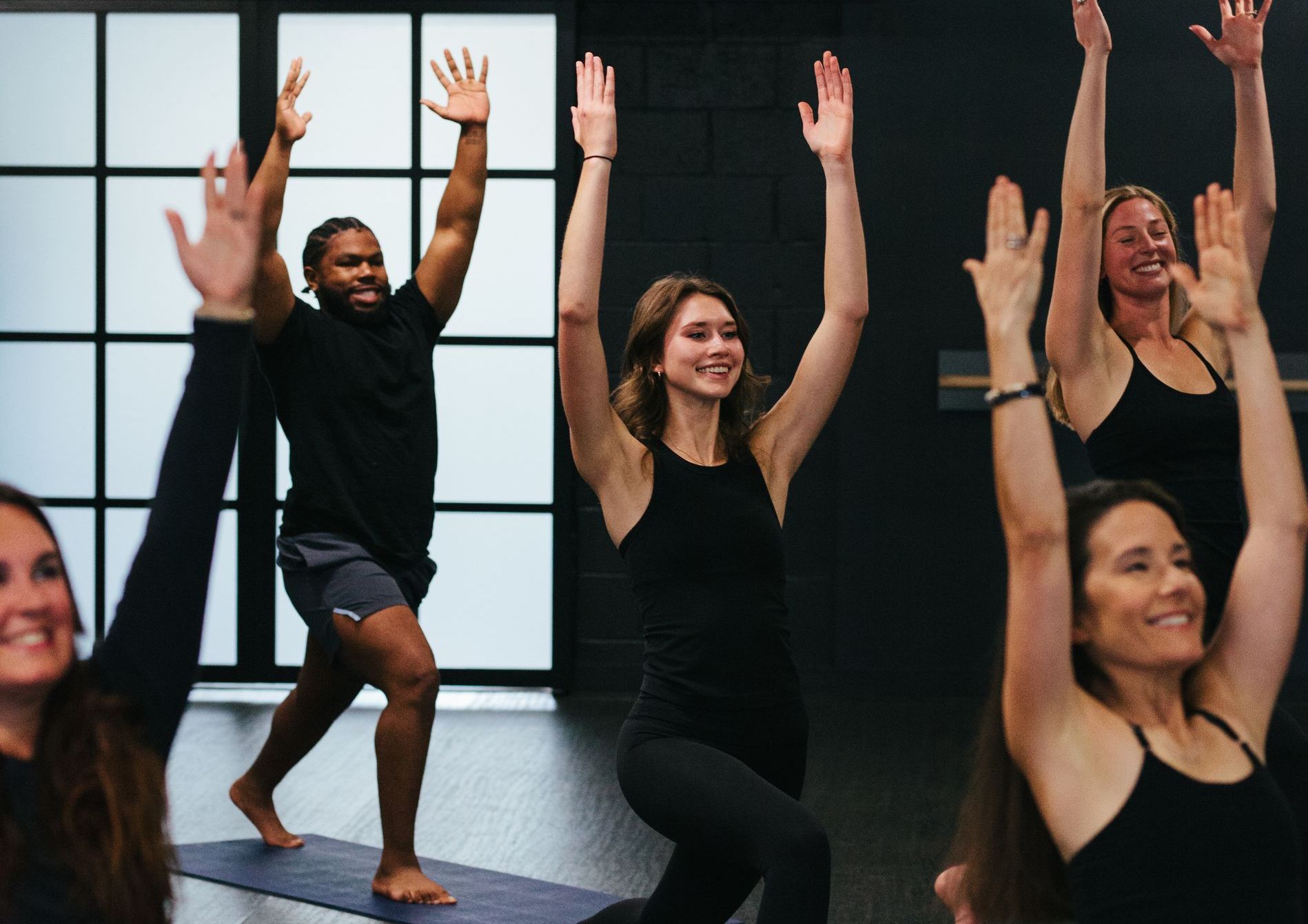 A group of four people practicing yoga in a studio with arms raised and smiling, wearing black athletic clothing.