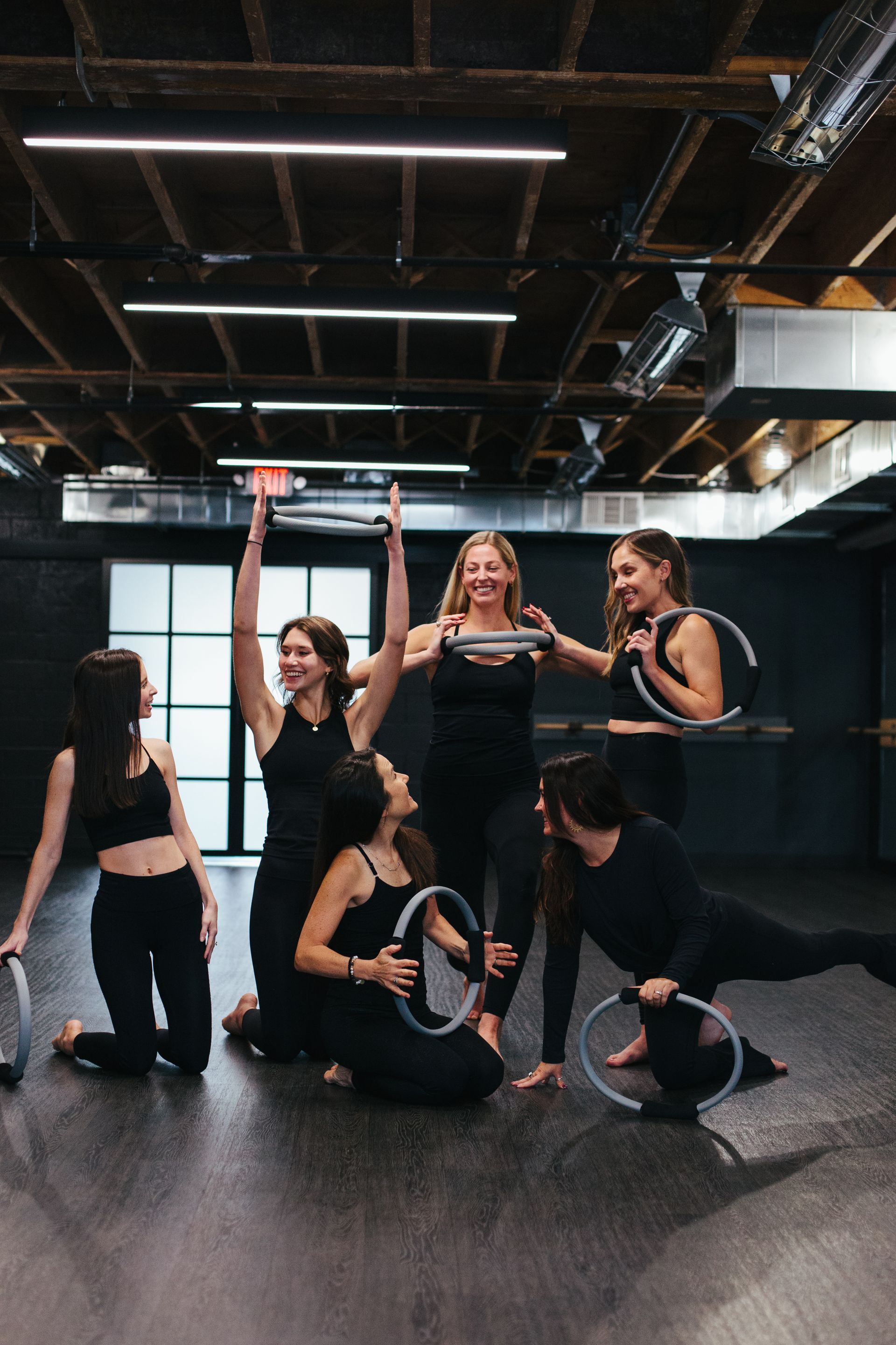 A group of six people in black workout attire pose with pilates rings in a studio with a dark ceiling and back wall.