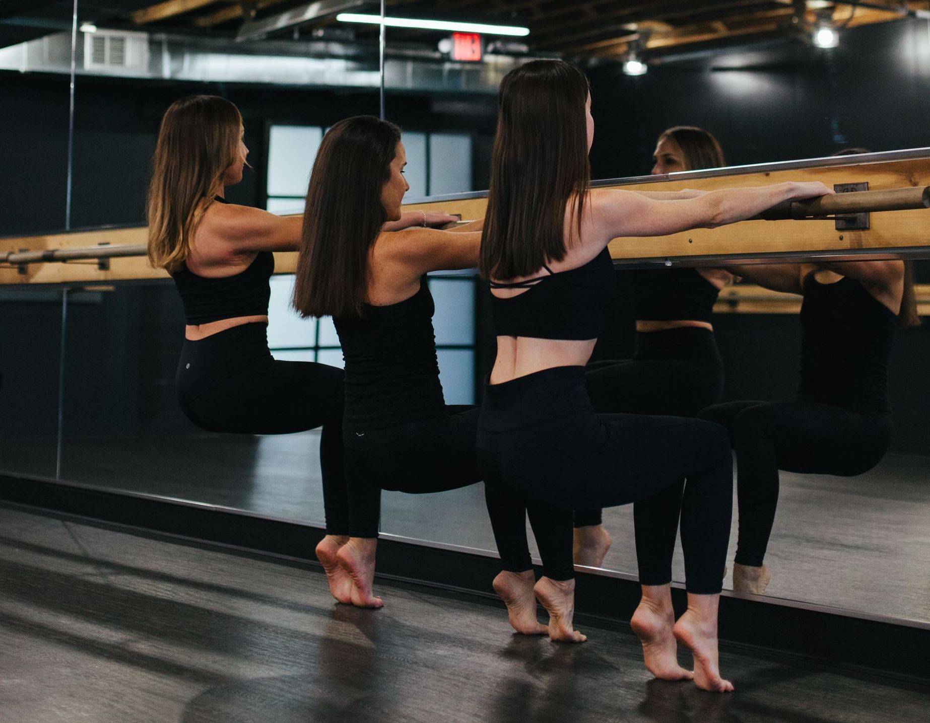 Two people in black activewear performing a barre squat exercise while holding onto a wooden studio barre.