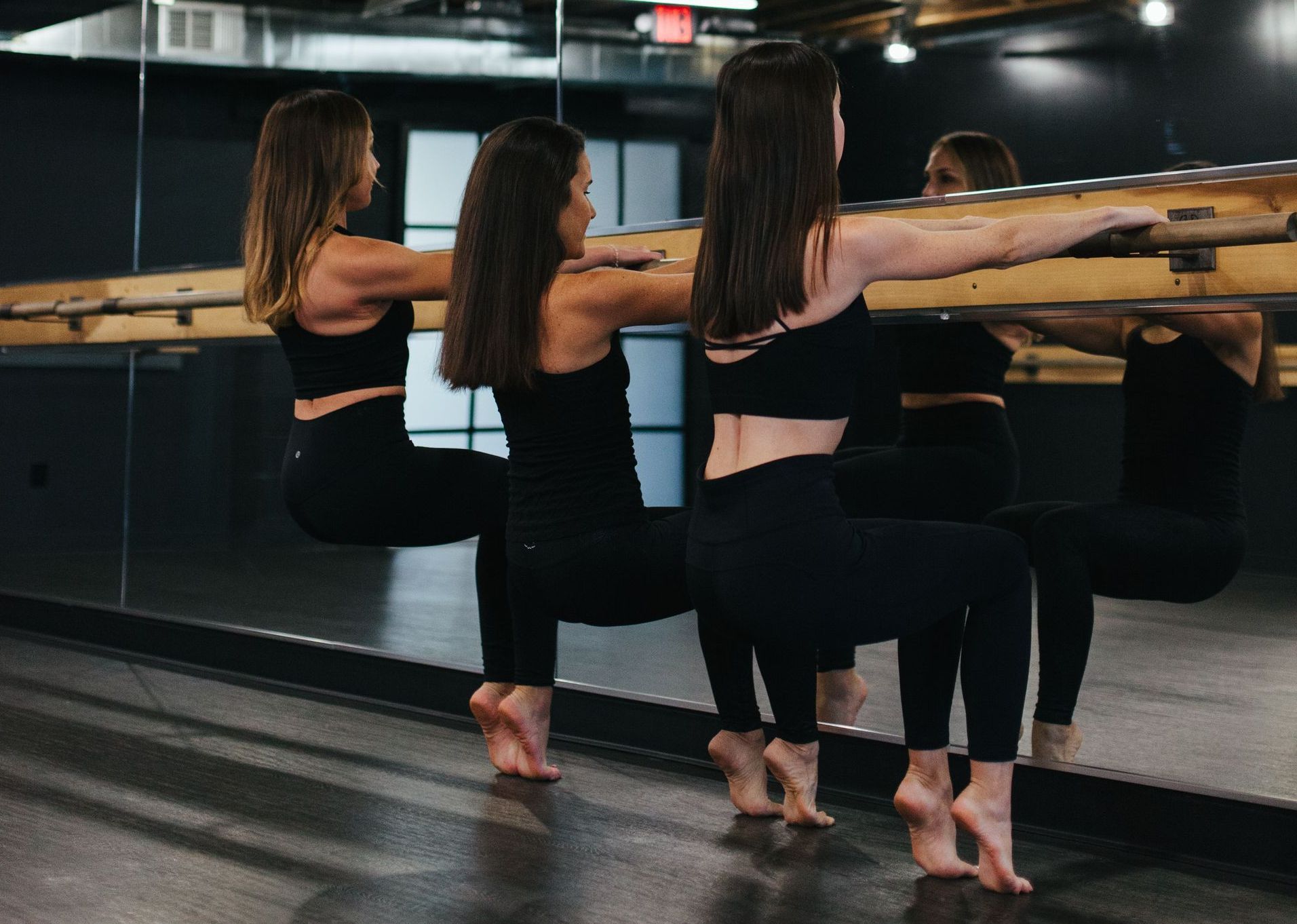Three people in black athletic wear perform a ballet barre squat on their tiptoes in a studio with a large mirror.