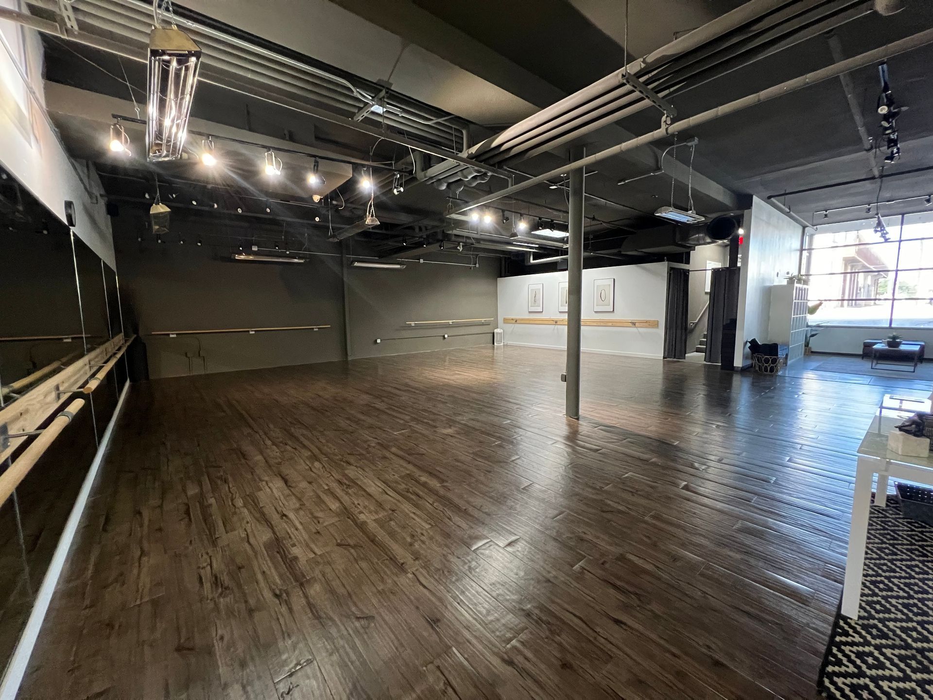 Empty dance studio with dark wood floors, wall-mounted ballet barres, mirrored walls, and exposed industrial ceiling.