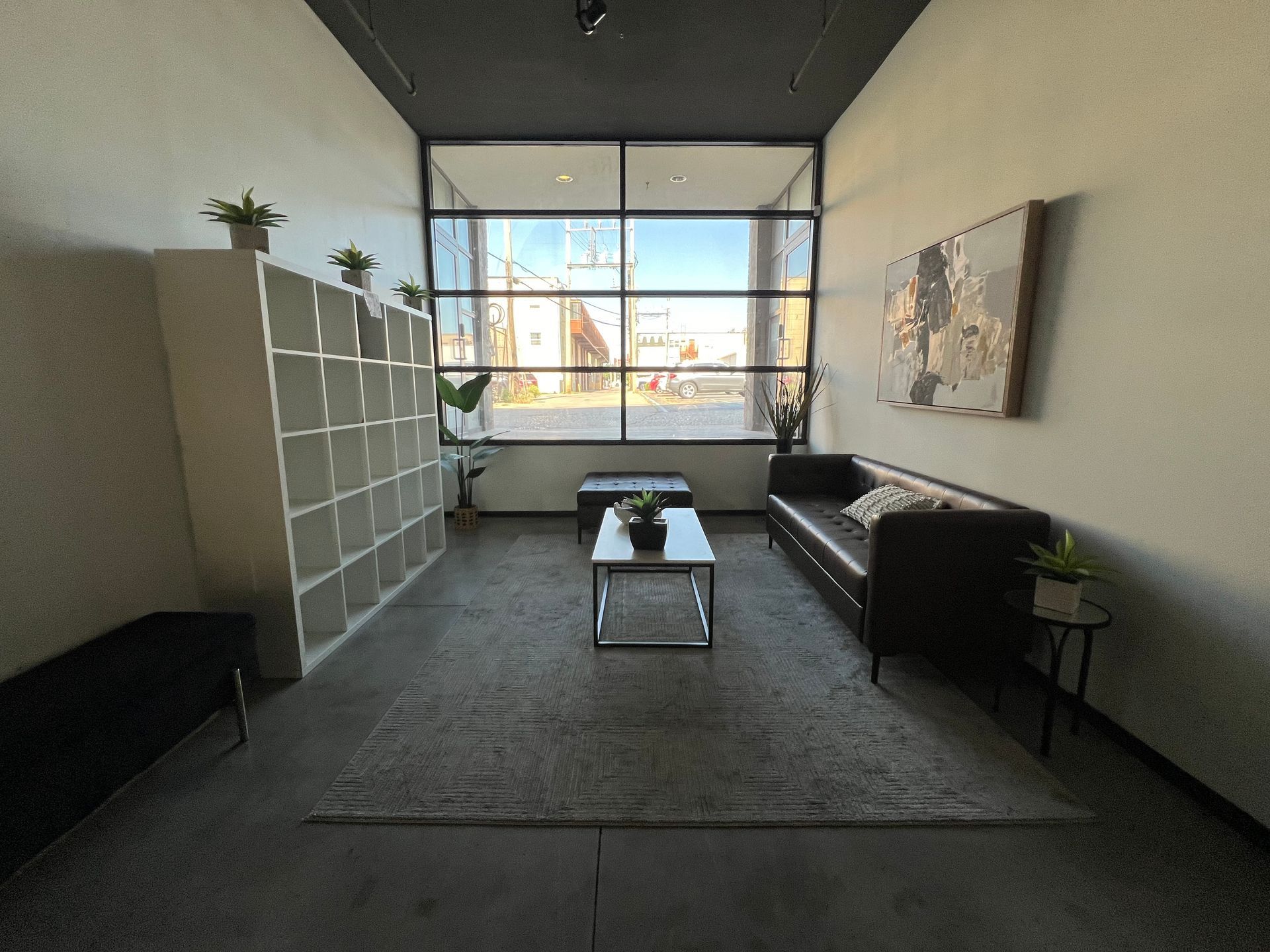 A modern waiting area with a grid shelving unit, black sofa, central coffee table, and a large window facing the street.