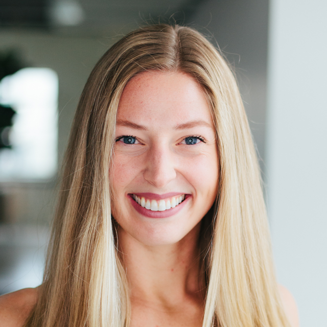 A smiling person with long blonde hair facing forward in a brightly lit indoor space.