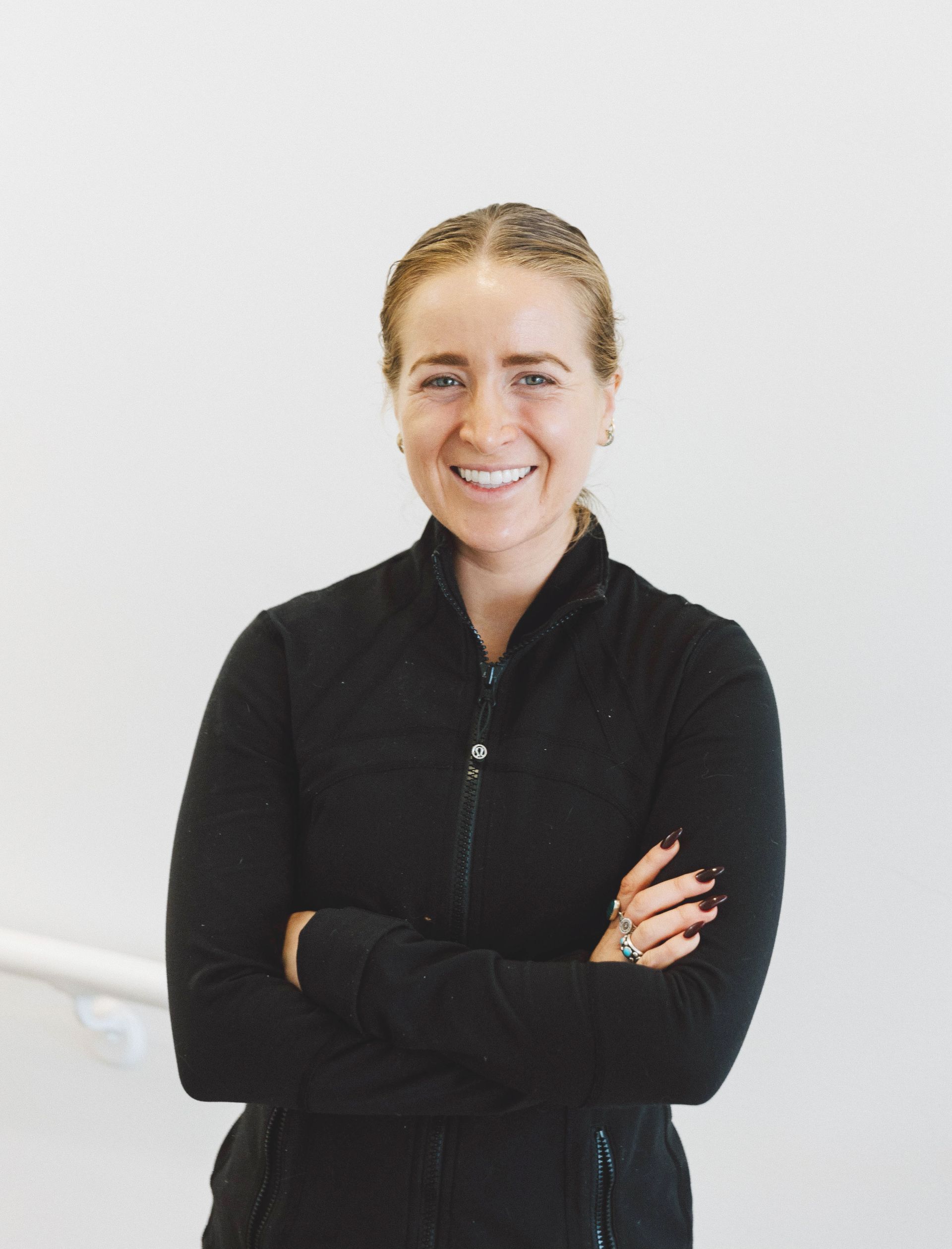 A smiling person with pulled-back hair wearing a black long-sleeve zip-up top stands with their arms crossed.