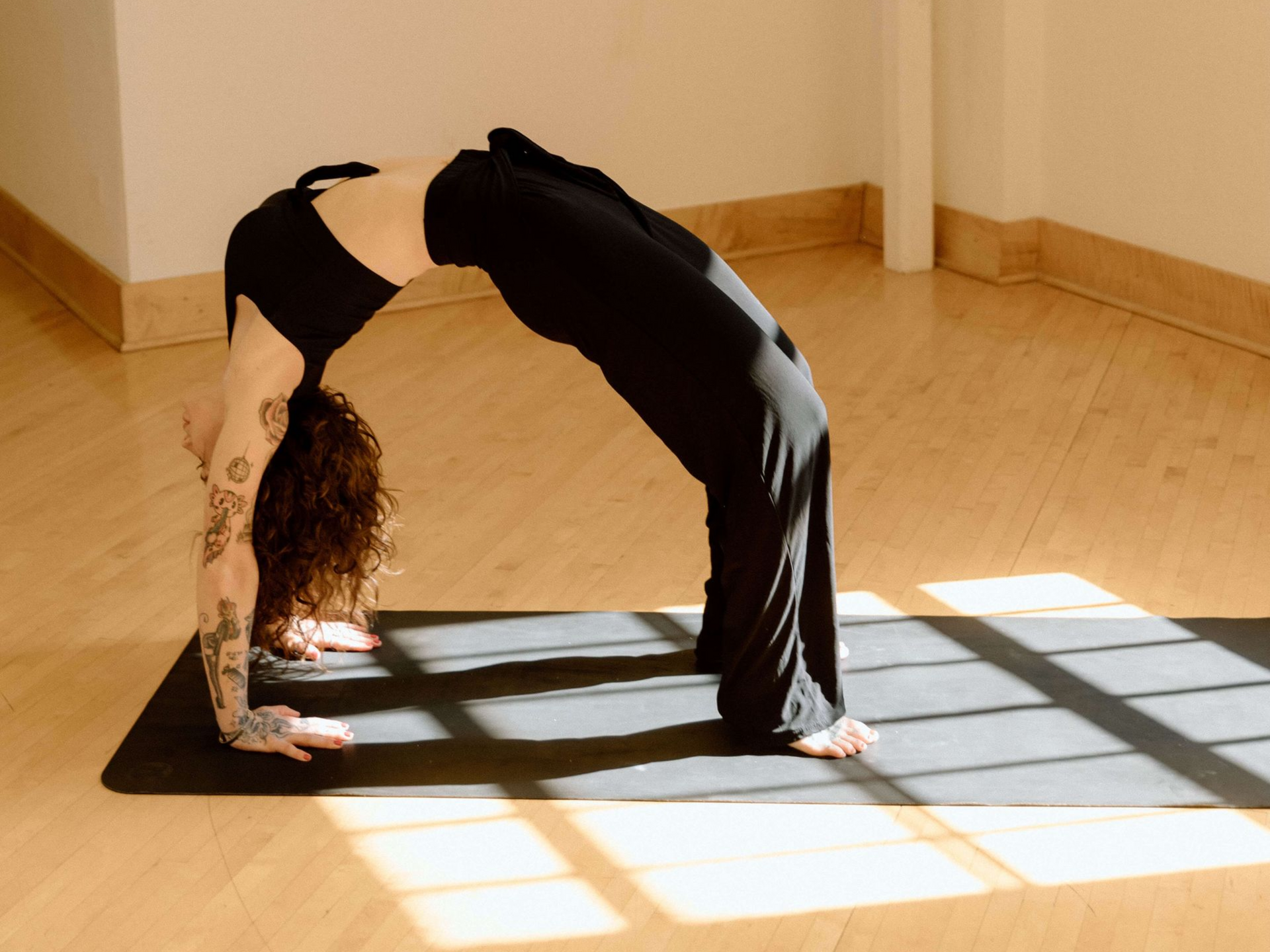 A person with tattooed arms performs a wheel yoga pose on a black mat in a sunlit room with wooden floors.