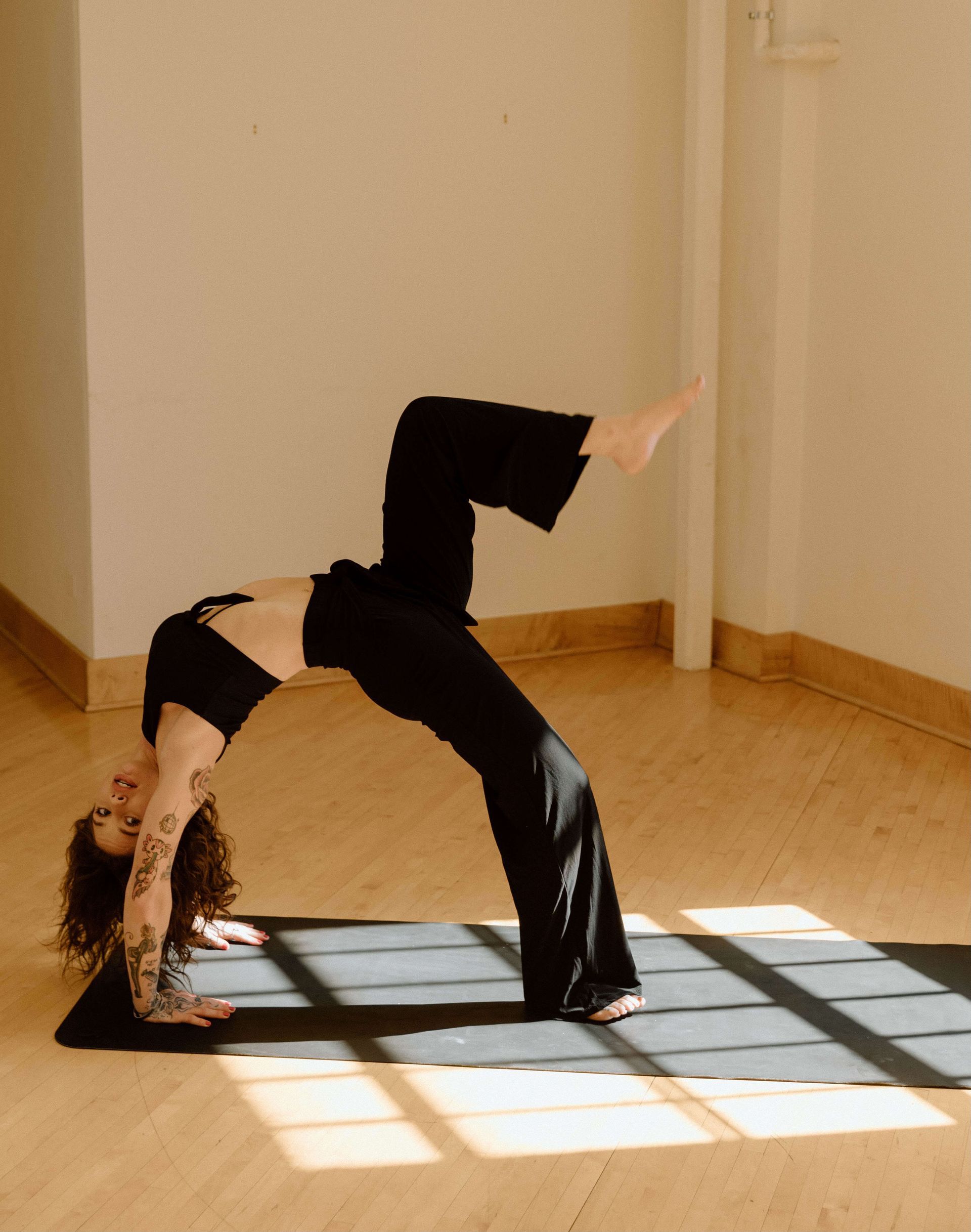 A person with tattoos practices a one-legged wheel yoga pose on a black mat in a bright, minimalist room.