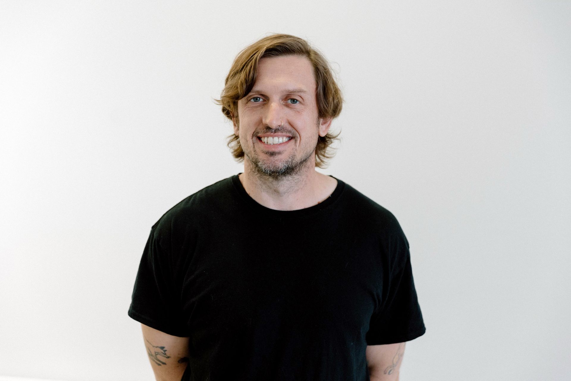 A person with shoulder-length wavy hair and a light beard smiles against a plain white background, wearing a black shirt.