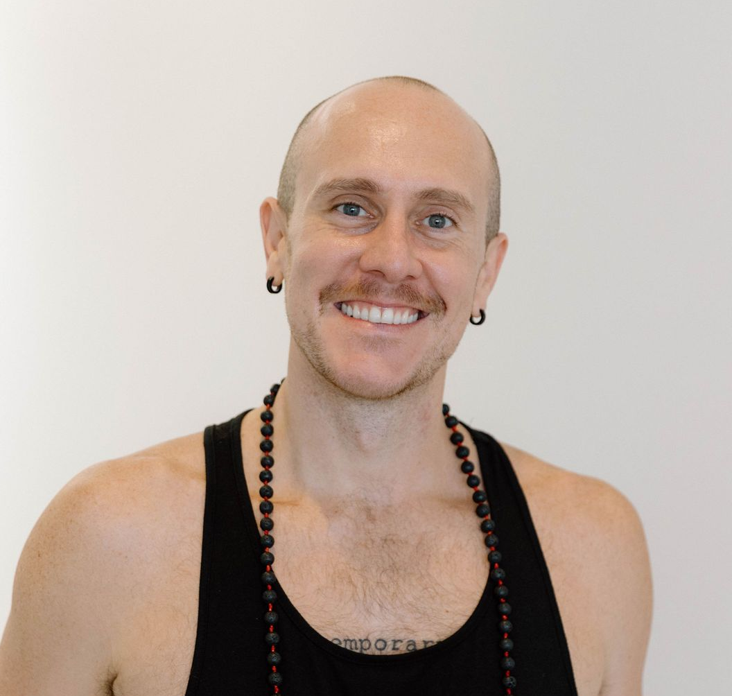 A person with a shaved head and mustache wearing a black tank top and bead necklace smiles against a white background.