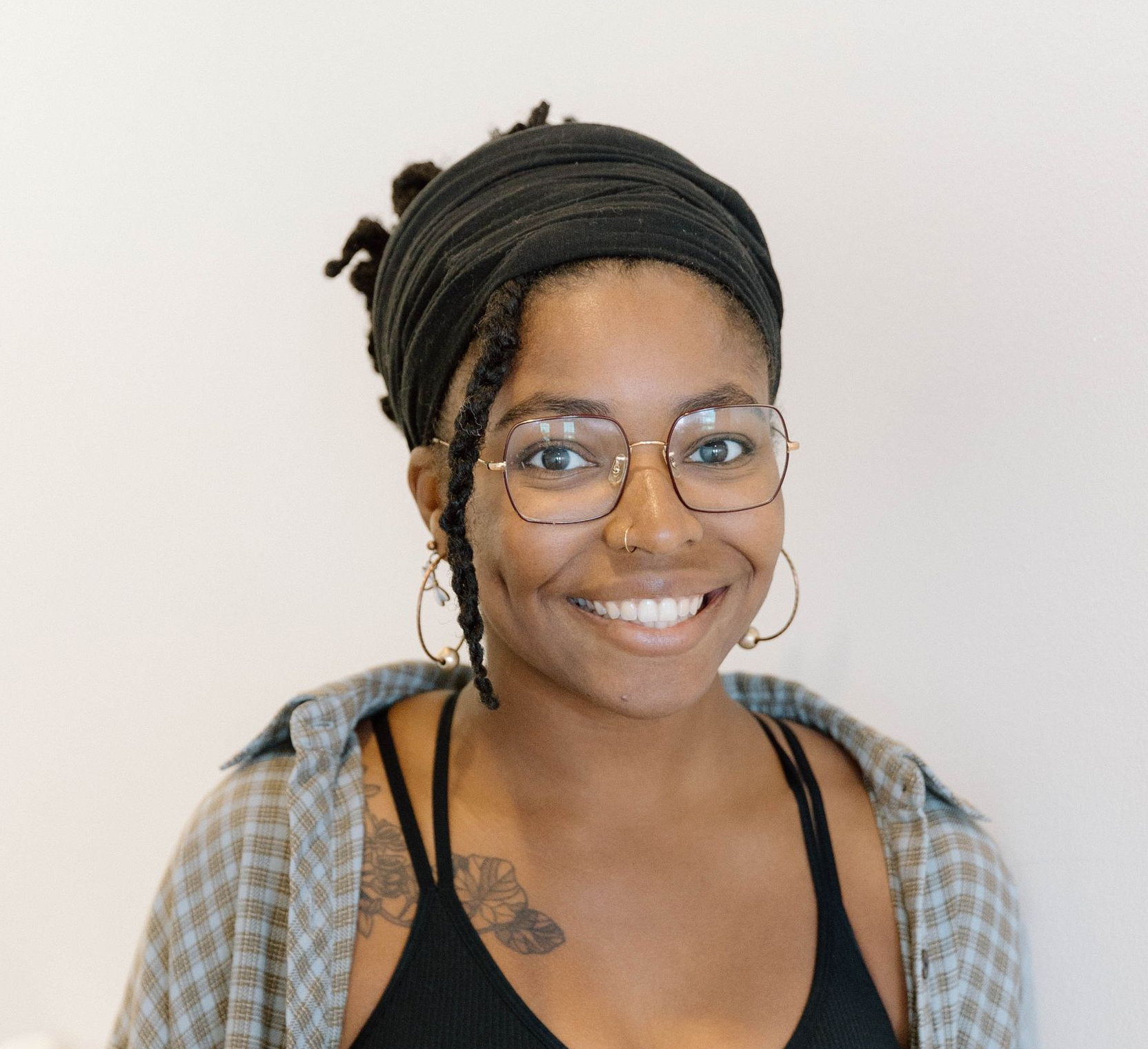A smiling person with dark hair, wearing a black headband, glasses, and gold hoop earrings, against a white background.