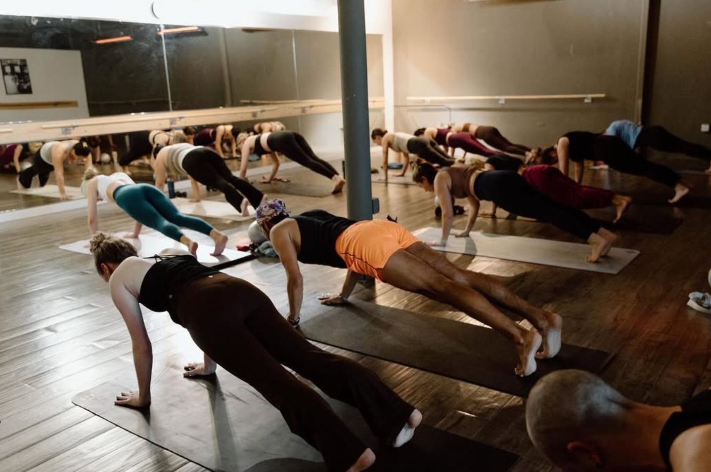 A group of people in a yoga studio hold a plank pose on exercise mats, reflected in a large wall mirror.