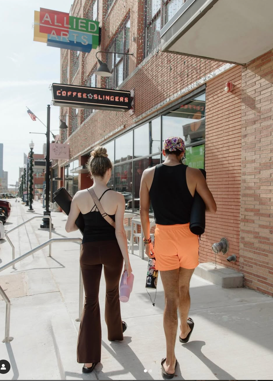 Two people walk down a city sidewalk toward an Allied Arts building, carrying yoga mats and wearing workout attire.