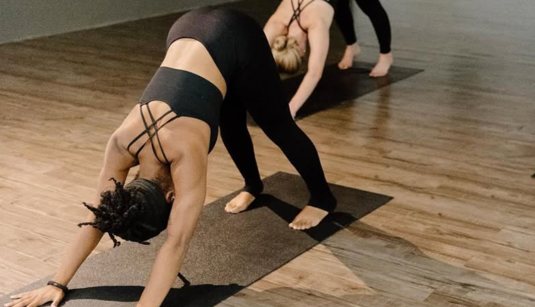 Two people in black athletic wear perform the downward-facing dog yoga pose on mats in a studio with wooden floors.