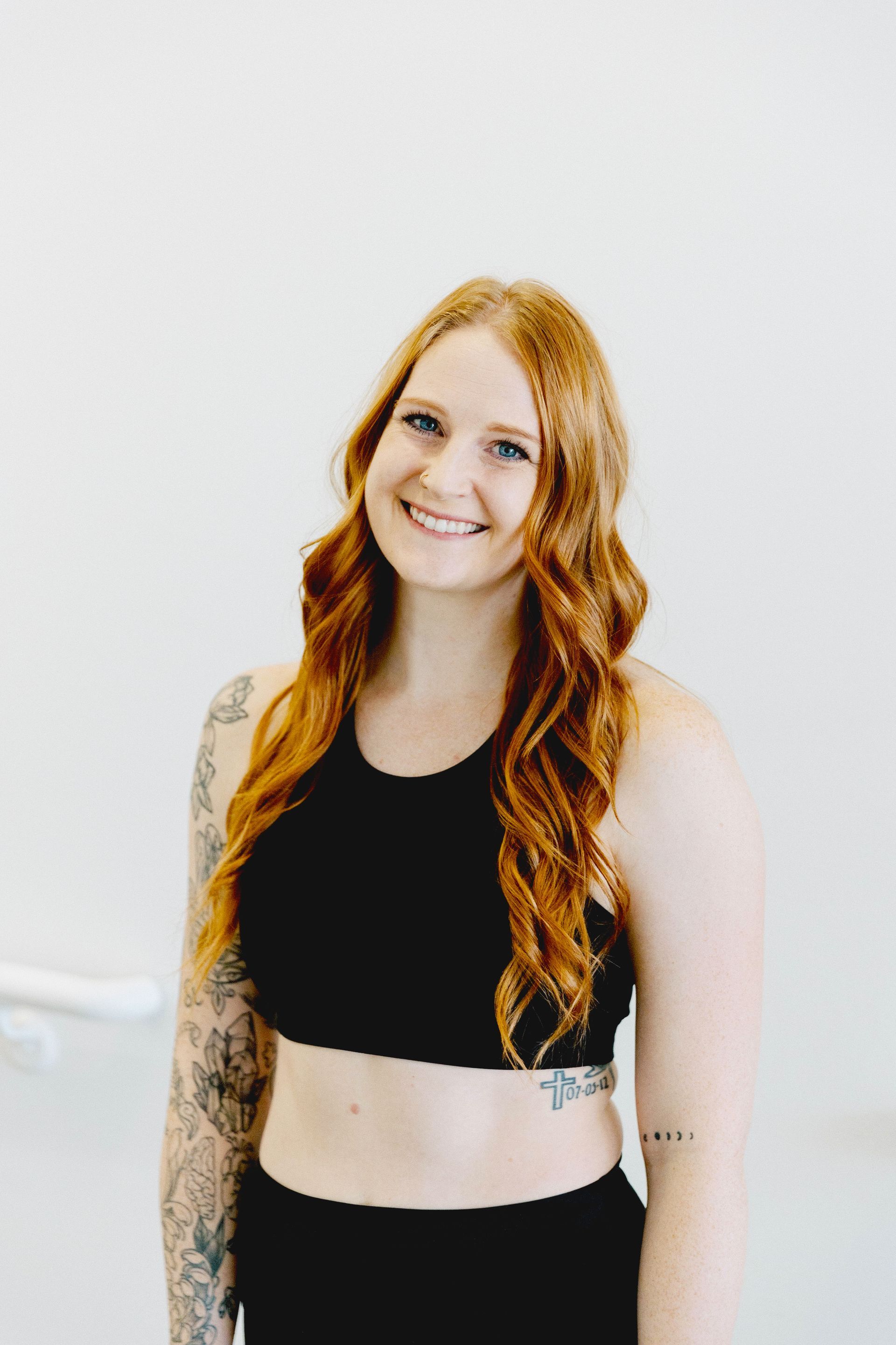 A person with red hair and arm tattoos smiling in a black sports top against a plain white background.