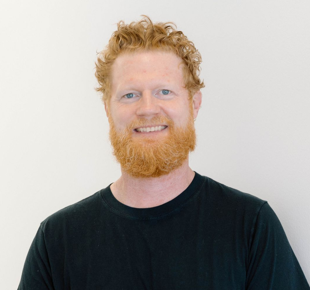 A person with curly reddish-blonde hair and a matching beard, wearing a black t-shirt, smiles against a plain background.