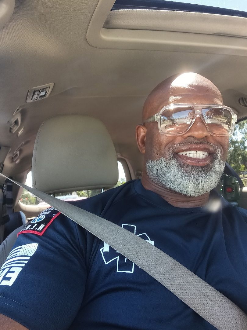 Smiling man wearing glasses and a seat belt in a car, sunlight on his face.
