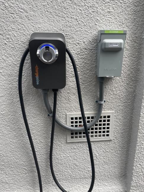 Wall-mounted electronic control box with knob and connected cable beside a gray vent on a textured wall