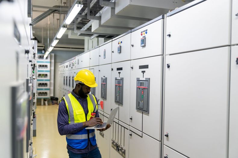 Worker in a yellow hard hat inspecting electrical panels in a bright industrial control room.
