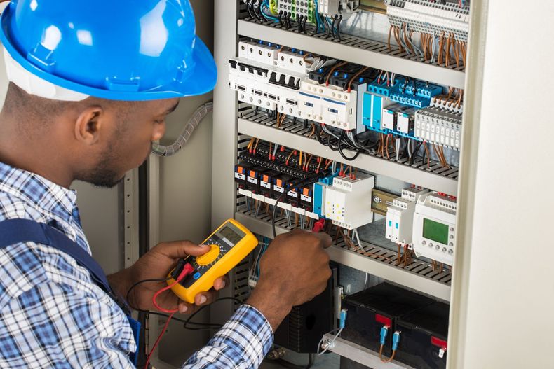 Technician in blue hard hat testing wiring inside an electrical control panel with a multimeter