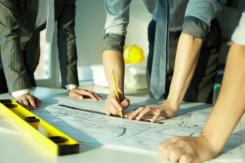 Persone che lavorano su un progetto con una livella, una matita e dei piani su un tavolo bianco.
