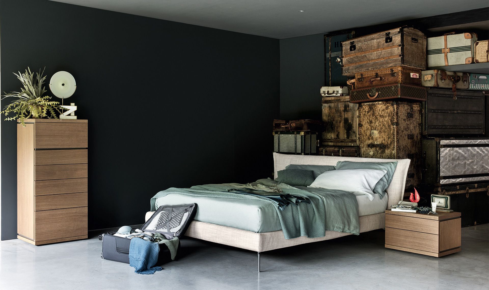Camera da letto con letto, cassettiera, comodino, valigie e bauli vintage impilati contro pareti scure.