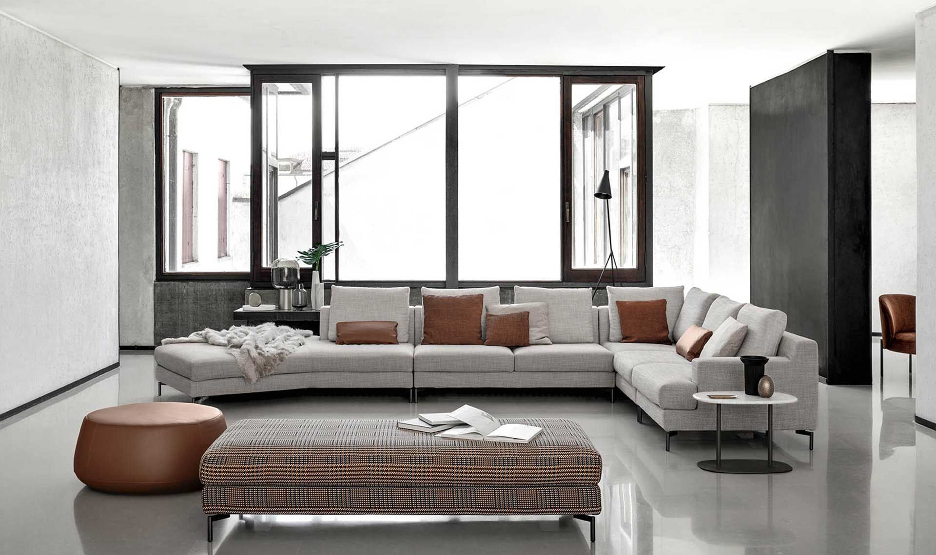 Soggiorno moderno con divano componibile, pouf marrone e ampie finestre.