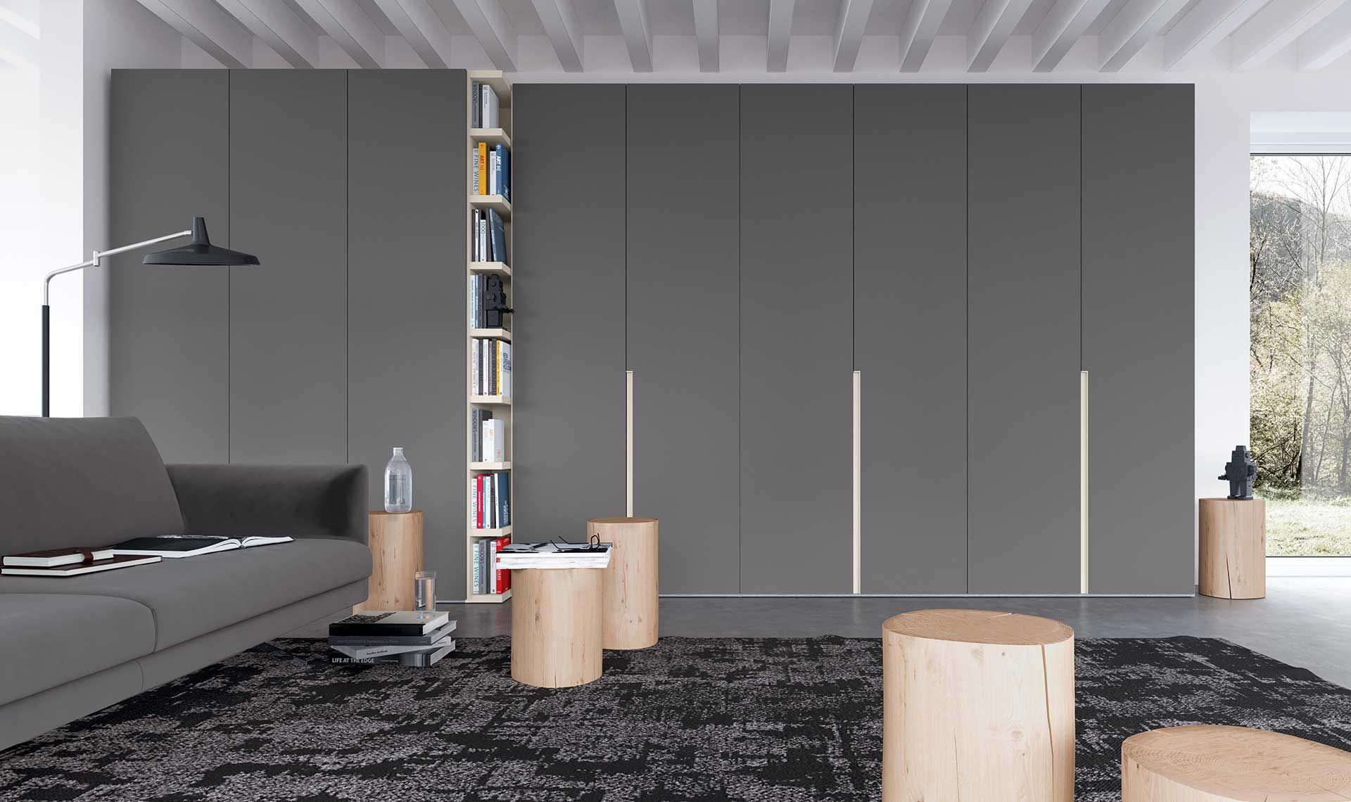 Soggiorno con armadi grigio scuro, libreria, divano grigio e sgabelli in legno su un tappeto a fantasia.