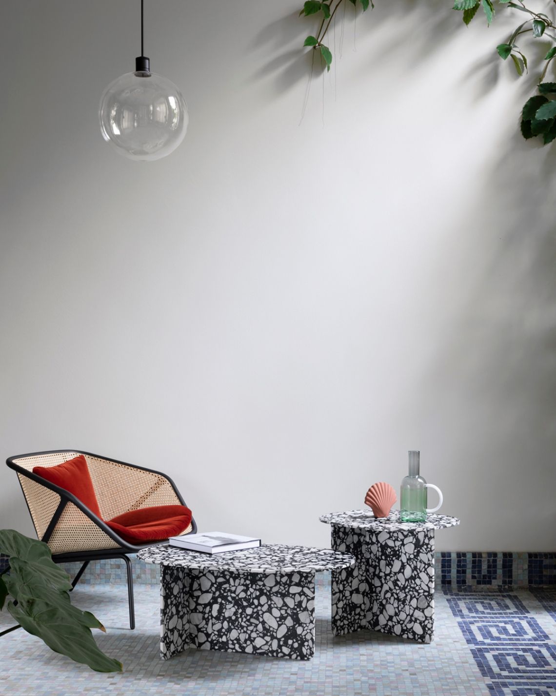 Una moderna area salotto all'aperto. Sedia con cuscino rosso, tavoli in terrazzo, lampada a globo, verde, parete bianca.