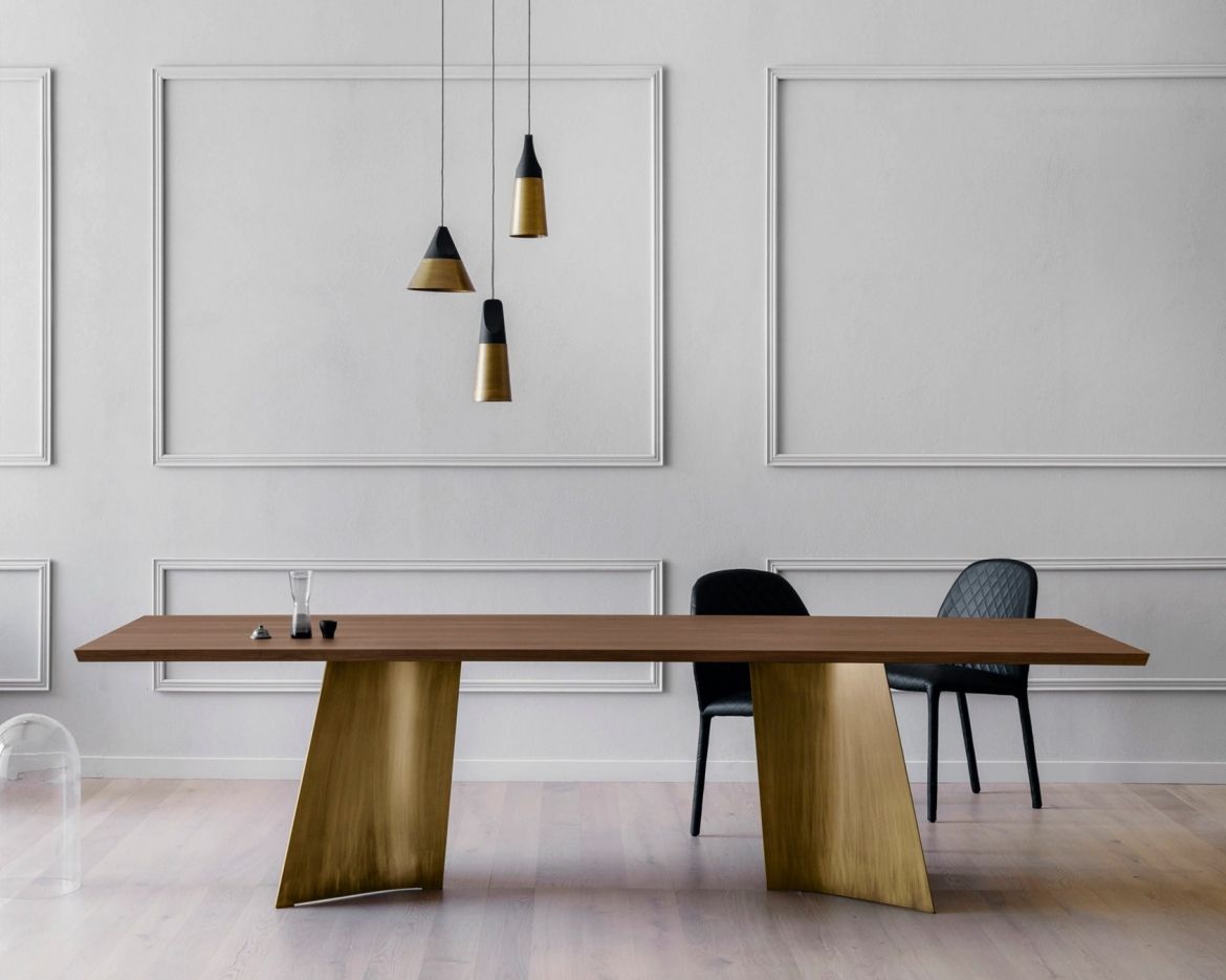 Sala da pranzo con un lungo tavolo marrone su supporti dorati, due sedie nere e lampade a sospensione.