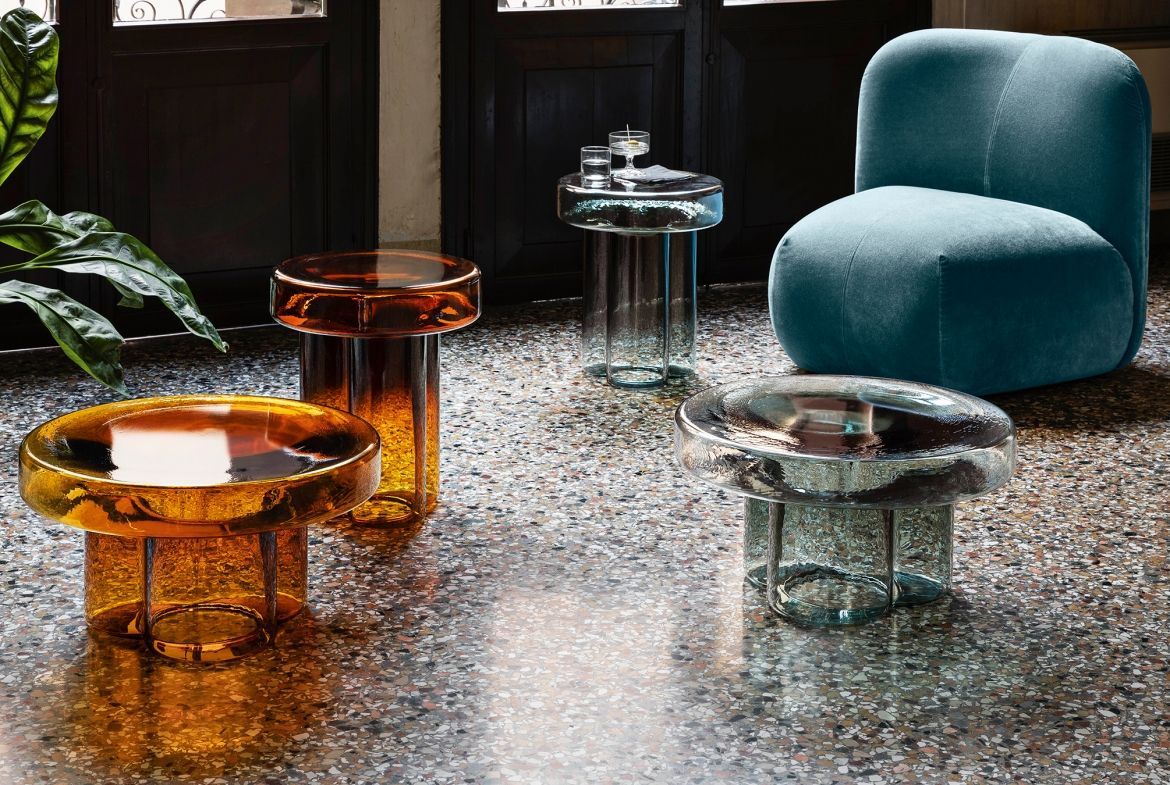 Tavolini laterali in vetro color ambra e verde acqua, su un pavimento punteggiato, con una poltrona verde acqua sullo sfondo.