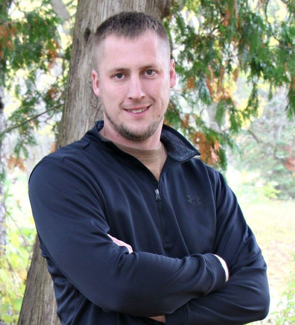 A man in a black jacket is standing in front of a tree with his arms crossed