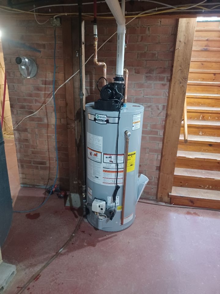 A water heater is sitting in a basement next to a brick wall.