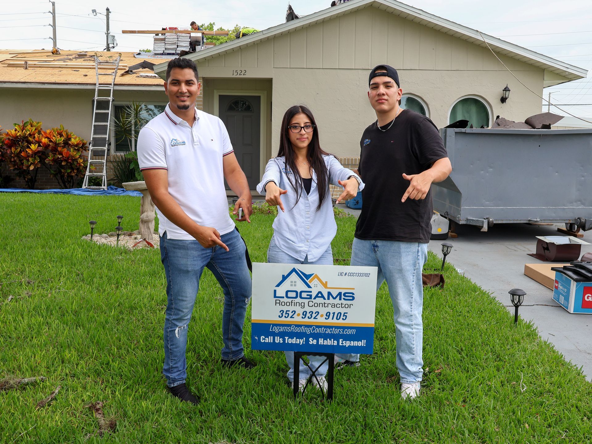 Three people stand in front of a house. They point at a sign advertising roofing services. Construction in progress.